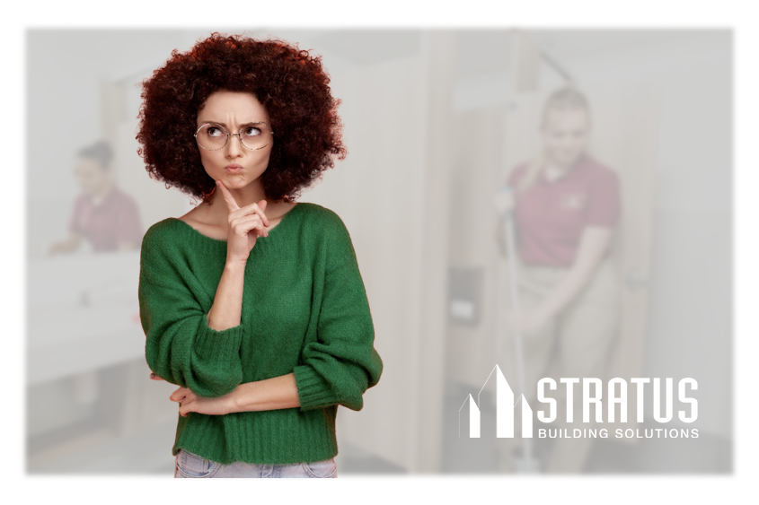 A woman wearing a green sweater and glasses while holding her hand to her chin with a curious expression and some people cleaning a restroom in the background.