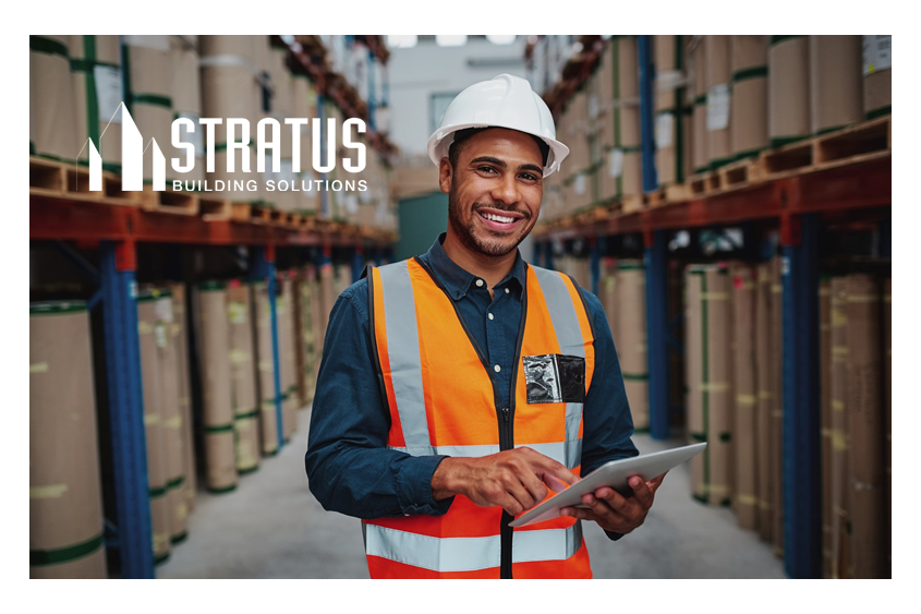 A smiling man in a hard hat and safety vest holds a tablet and stands in a warehouse full of shelves of rolled up materials 