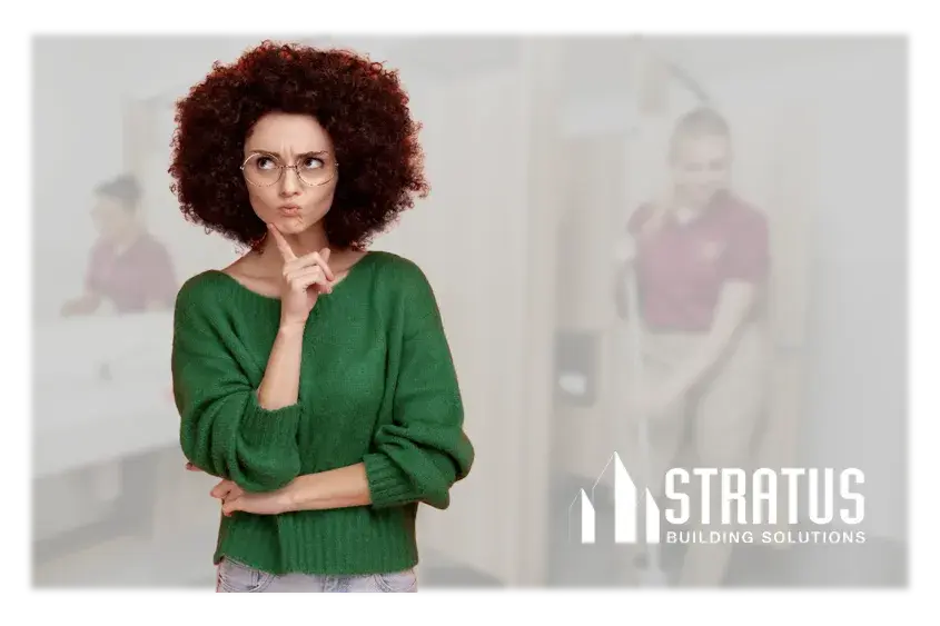 A woman wearing a green sweater and glasses while holding her hand to her chin with a curious expression and some people cleaning a restroom in the background.